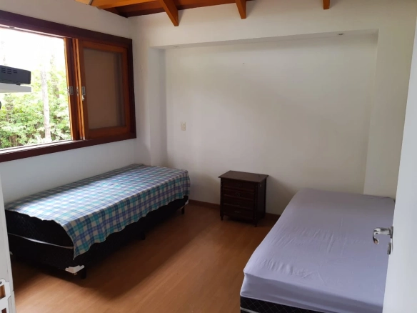 Imagem de um quarto bem iluminado, com duas camas de solteiro, uma mesa de cabeceira e um grande painel de janelas que permitem a entrada de luz natural. CT Rezende