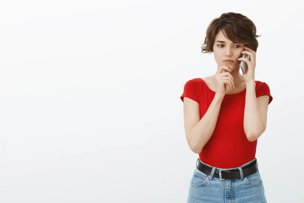 Mulher jovem preocupada usando um telefone celular, vestindo camiseta vermelha e jeans, pensando com expressão de dúvida contra fundo branco.