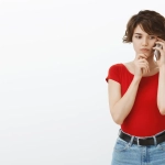 Mulher jovem preocupada usando um telefone celular, vestindo camiseta vermelha e jeans, pensando com expressão de dúvida contra fundo branco.