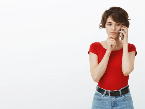 Mulher jovem preocupada usando um telefone celular, vestindo camiseta vermelha e jeans, pensando com expressão de dúvida contra fundo branco.